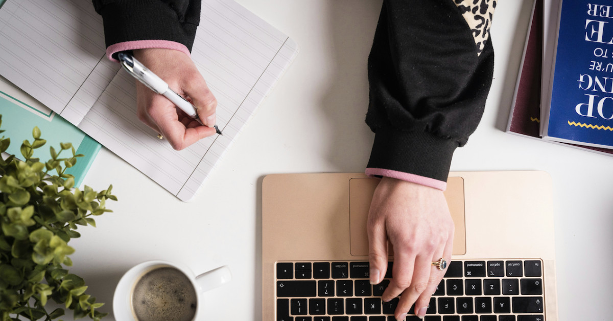 Person writing in a notebook next to a laptop on a desk with coffee and a plant, photographed from above.