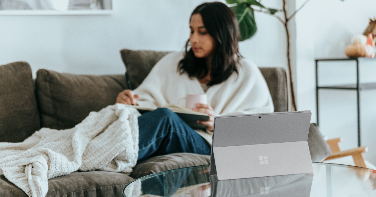Woman on the couch with a blanket, a notebook, and a cup of coffee, with a laptop on the coffee table in front of her.