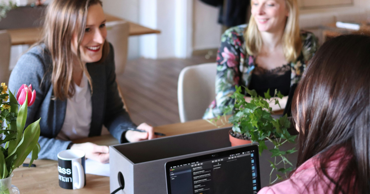 Three women in conversation at a work table with laptop, coffee and flowers, in an informal office environment.