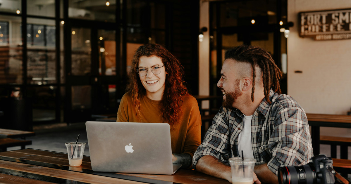 Two people laughing together at a laptop in a casual café, with coffee and a camera on the table.